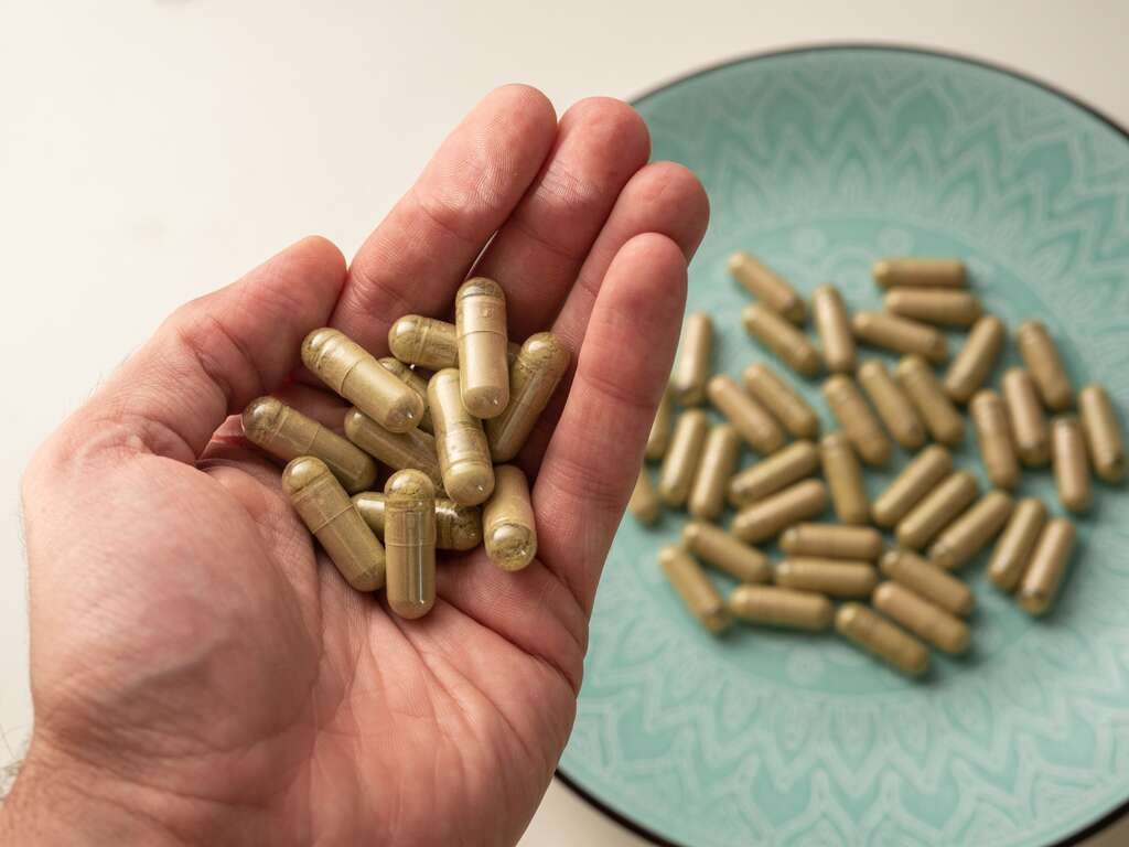 Hand met kratomcapsules en nog meer capsules op een blauwgroen bord